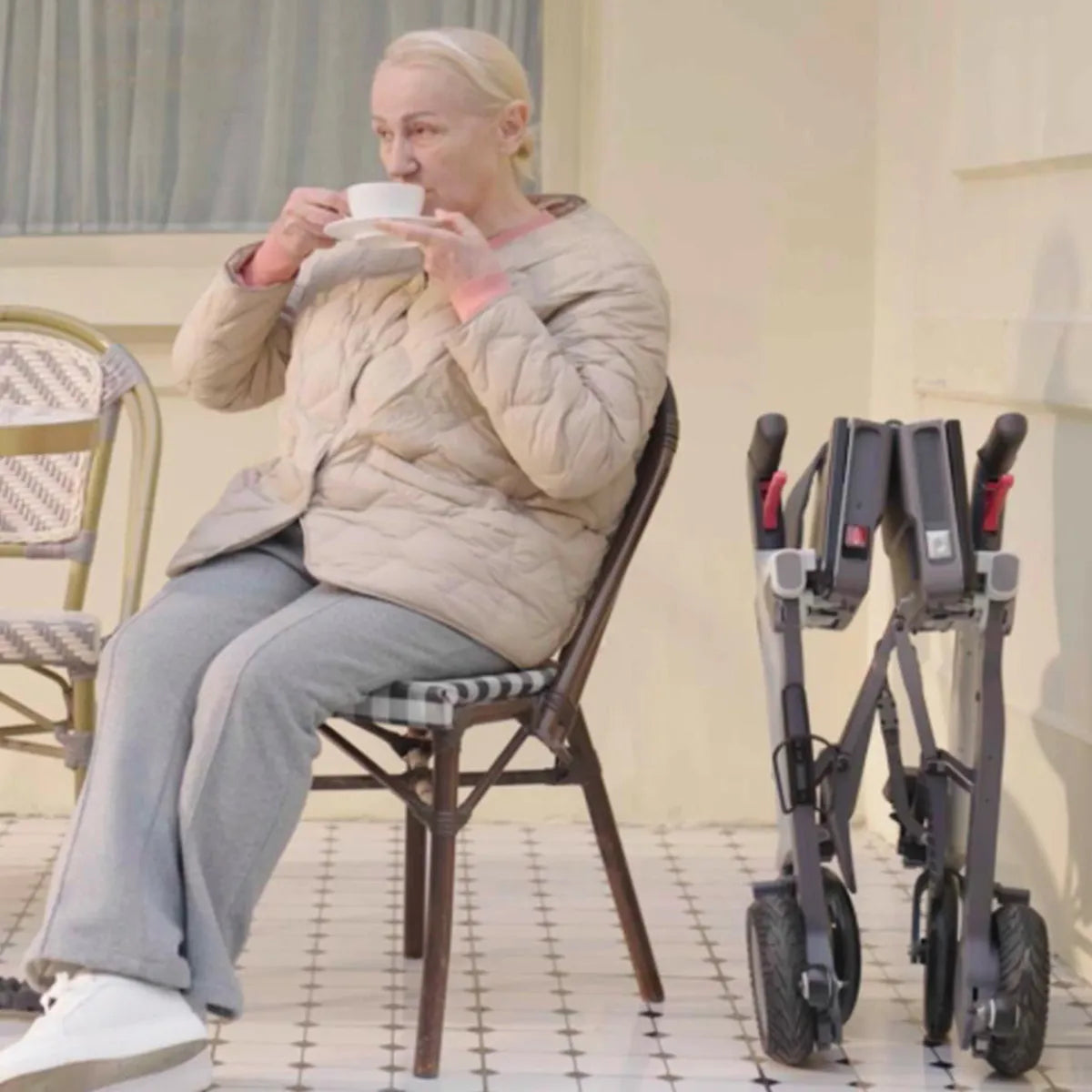 Ältere Frau sitzt beim Kaffeetrinken, neben ihr steht der zusammengeklappte Roamate – kompakter elektrischer Rollstuhl für Alltag und Reisen.
