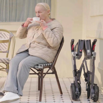 Ältere Frau sitzt beim Kaffeetrinken, neben ihr steht der zusammengeklappte Roamate – kompakter elektrischer Rollstuhl für Alltag und Reisen.